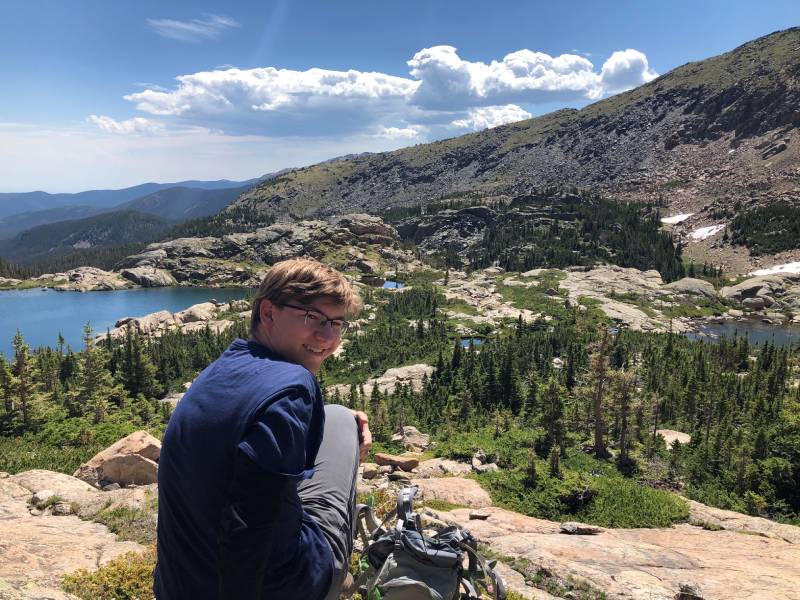 A picture of Carter sitting on a rocky overlook with a rocky mountain and lake in the background